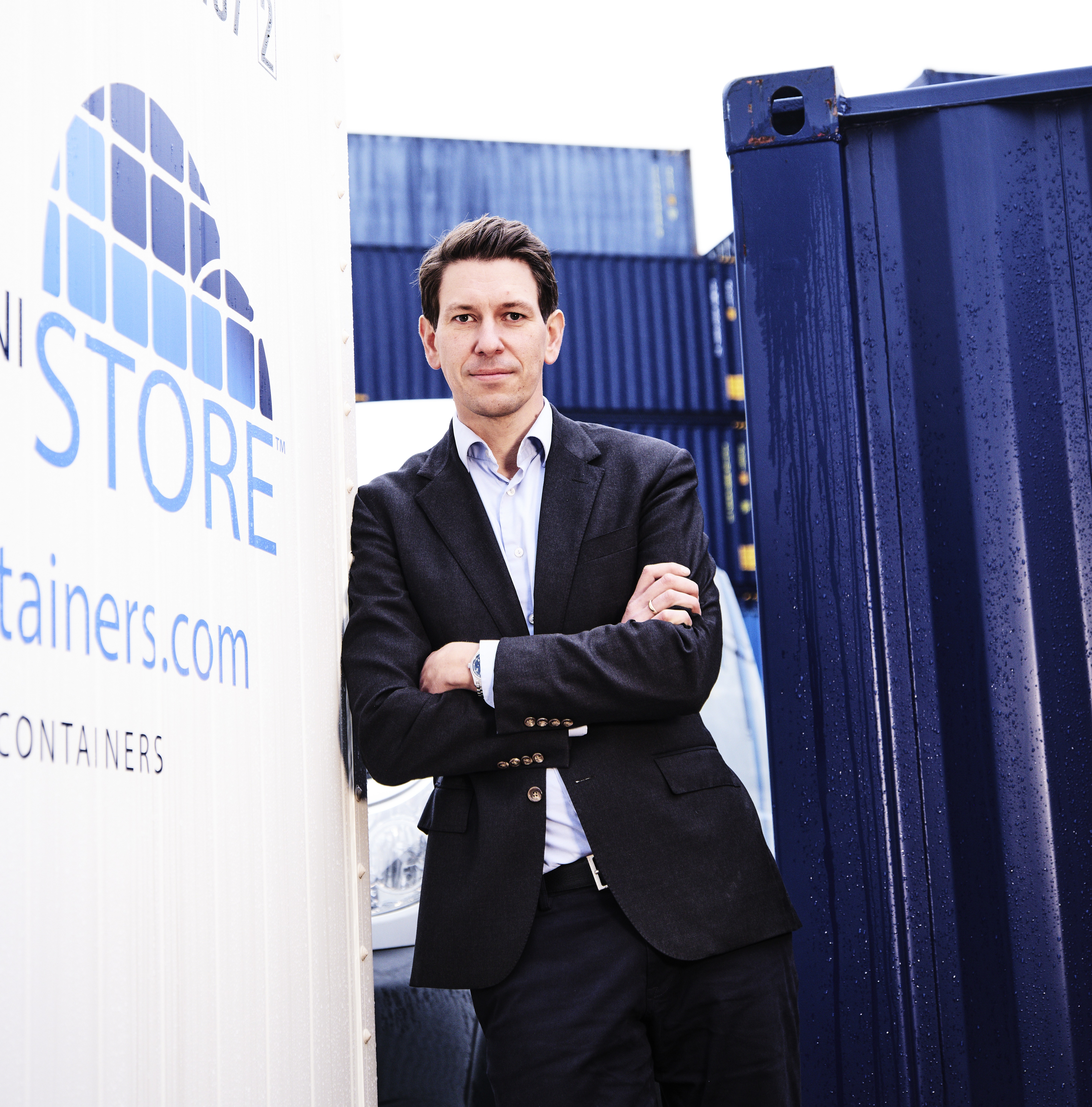 Professional man in dark blazer and white shirt standing confidently with arms crossed in front of blue shipping containers and company signage Headshot