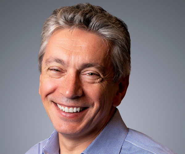 Professional headshot of smiling middle-aged man in light blue striped shirt against gray background