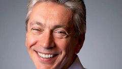Professional headshot of smiling middle-aged man in light blue striped shirt against gray background
