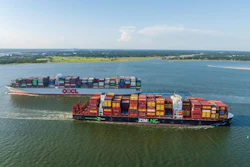 Two container cargo ships loaded with colorful shipping containers sailing in coastal waters with green shoreline in background