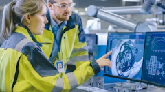 Two engineers in yellow safety vests reviewing 3D CAD design on computer monitor in industrial facility