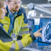 Two engineers in yellow safety vests reviewing 3D CAD design on computer monitor in industrial facility