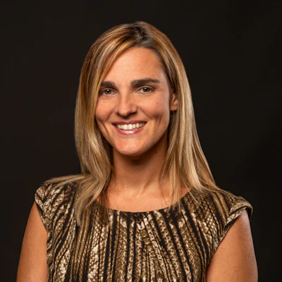 Professional headshot of a smiling woman with long blonde hair wearing a patterned bronze-toned top against a dark background