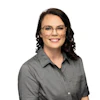 Professional headshot of a woman wearing glasses and a gray button-up shirt, smiling at camera