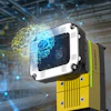 Industrial vision camera with white and black housing mounted on yellow equipment in a smart factory warehouse with digital network visualization