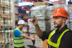 Warehouse worker in orange hard hat and safety vest using tablet with digital business analytics icons overlay