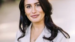 Professional headshot of woman with dark wavy hair wearing light gray blazer