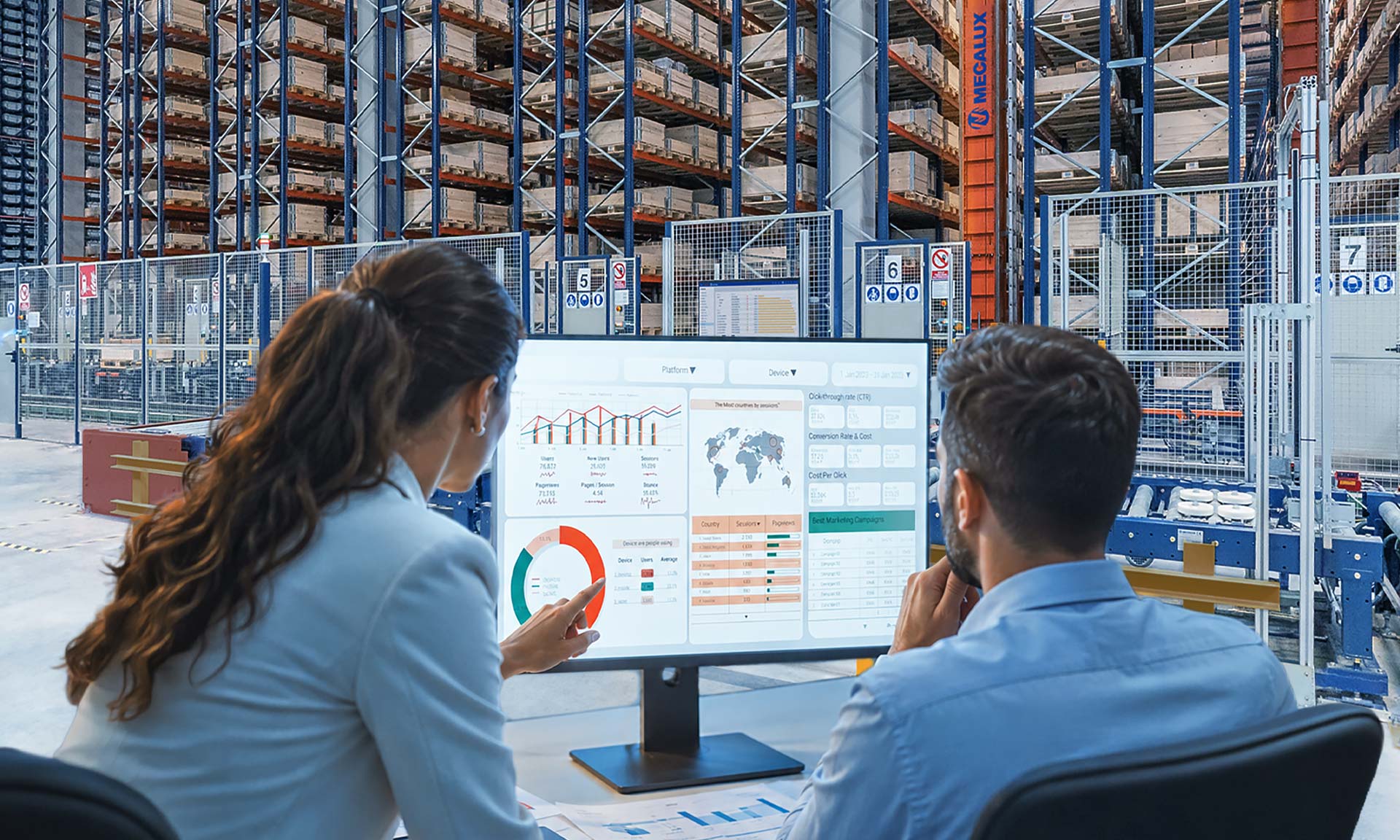 Two professionals reviewing analytics dashboards on a monitor in a warehouse with storage racks in the background