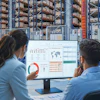 Two professionals reviewing analytics dashboards on a monitor in a warehouse with storage racks in the background