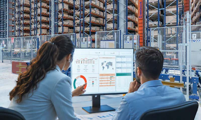 Two professionals reviewing analytics dashboards on a monitor in a warehouse with storage racks in the background