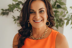 Professional headshot of woman in orange sleeveless top with gold chain necklace, smiling against green plant background