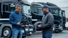 Two workers review documents at a logistics yard with commercial trucks in the background