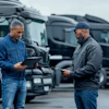 Two workers review documents at a logistics yard with commercial trucks in the background