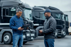 Two workers review documents at a logistics yard with commercial trucks in the background