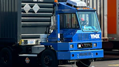 Blue industrial container handling truck with hydraulic crane lifting a dark shipping container in a logistics yard