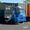 Blue industrial container handling truck with hydraulic crane lifting a dark shipping container in a logistics yard