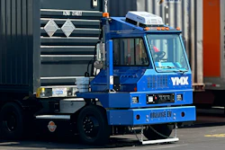 Blue industrial container handling truck with hydraulic crane lifting a dark shipping container in a logistics yard