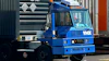 Blue industrial container handling truck with hydraulic crane lifting a dark shipping container in a logistics yard
