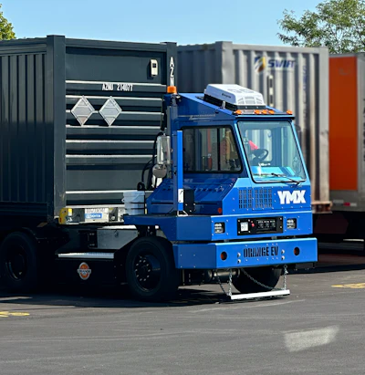Blue industrial container handling truck with hydraulic crane lifting a dark shipping container in a logistics yard