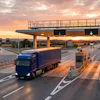 Trucks passing through highway toll booths at sunset with overpass and multiple lanes
