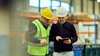 Two workers in hard hats reviewing data on a tablet in an industrial warehouse facility