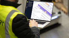 Worker in high-visibility vest using laptop displaying purple and orange data visualization interface in warehouse setting