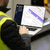 Worker in high-visibility vest using laptop displaying purple and orange data visualization interface in warehouse setting