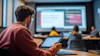 Student holding tablet in classroom watching presentation on large screen with peers seated at table