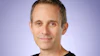Professional headshot of a middle-aged man wearing a black t-shirt against a purple gradient background