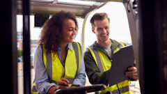 Two smiling workers in yellow safety vests reviewing documents on a tablet outdoors at a construction site