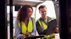Two smiling workers in yellow safety vests reviewing documents on a tablet outdoors at a construction site