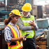 Two industrial workers in safety gear reviewing information on tablet in manufacturing facility