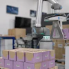 Collaborative robot arm sorting purple and brown cardboard boxes in a warehouse facility