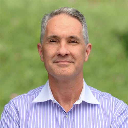 Professional headshot of a middle-aged man wearing a light purple striped shirt with white collar against a blurred green background Headshot