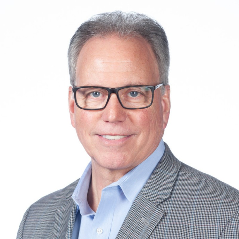 Professional headshot of a middle-aged man wearing glasses and a gray blazer with light blue shirt