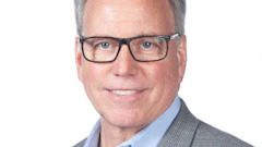 Professional headshot of a middle-aged man wearing glasses and a gray blazer with light blue shirt