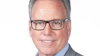 Professional headshot of a middle-aged man wearing glasses and a gray blazer with light blue shirt
