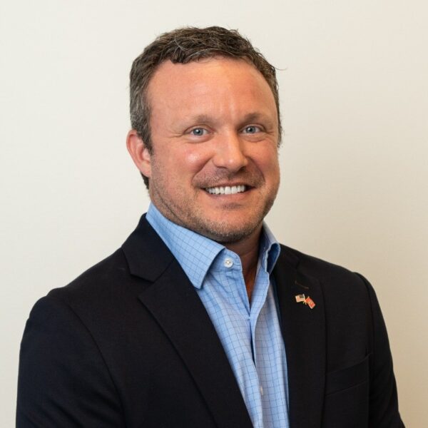 Professional headshot of a smiling man in dark blazer and blue checkered shirt with flag pin