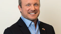 Professional headshot of a smiling man in dark blazer and blue checkered shirt with flag pin
