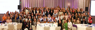 Large group of professional women and men in business attire standing together at a corporate conference or networking event with tables and stage backdrop.